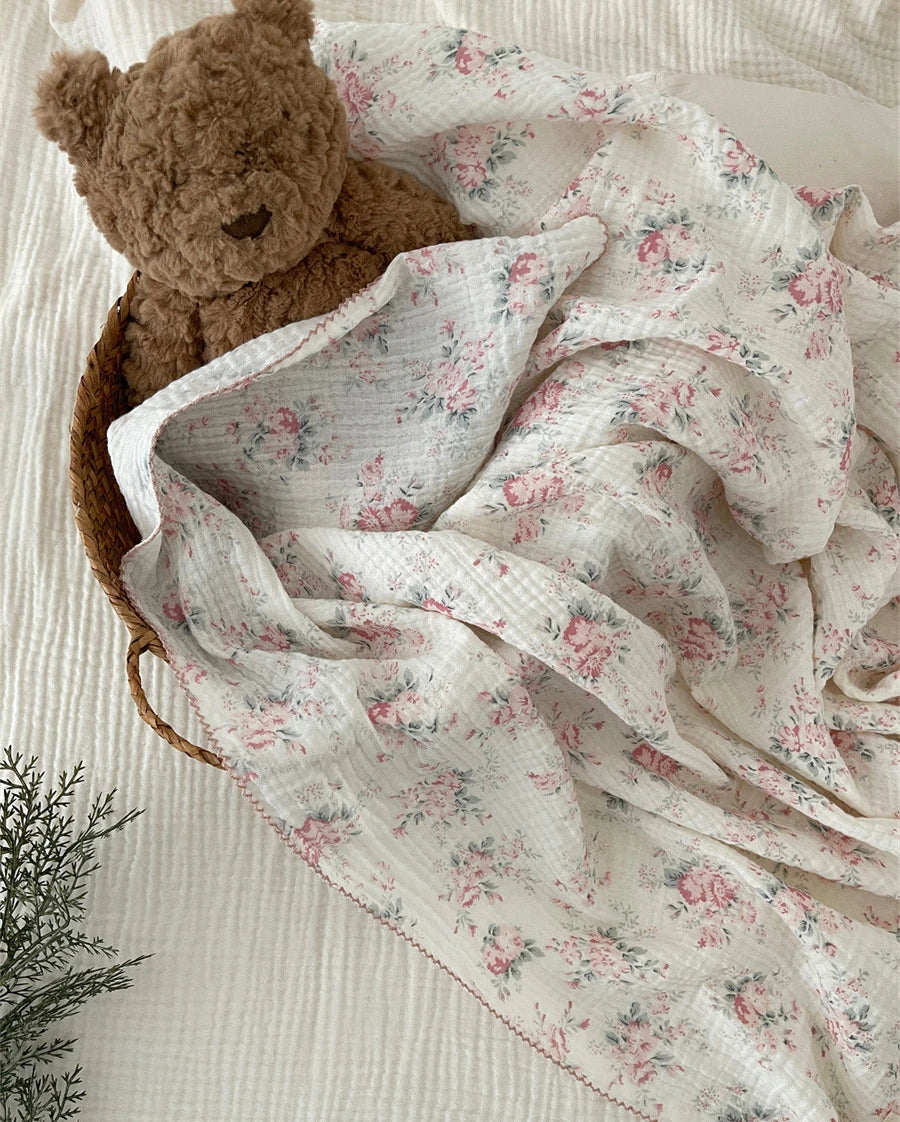 A soft white muslin swaddle blanket with a delicate floral print is draped next to a brown teddy bear.
