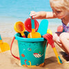 A young child plays with a teal plastic bucket filled with various orange and yellow beach sand tools on a sandy beach.