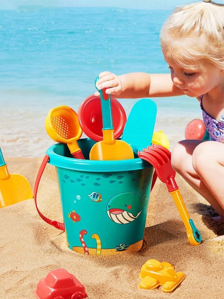 A young child plays with a teal plastic bucket filled with various orange and yellow beach sand tools on a sandy beach.