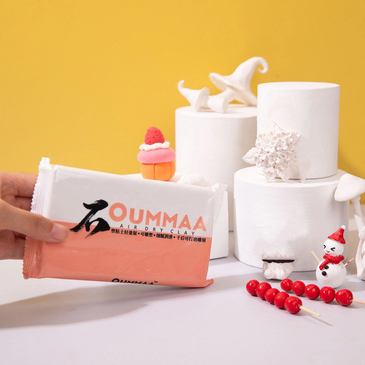 A hand holds a block of pink air-dry clay next to white sculpted figures on a yellow background.
