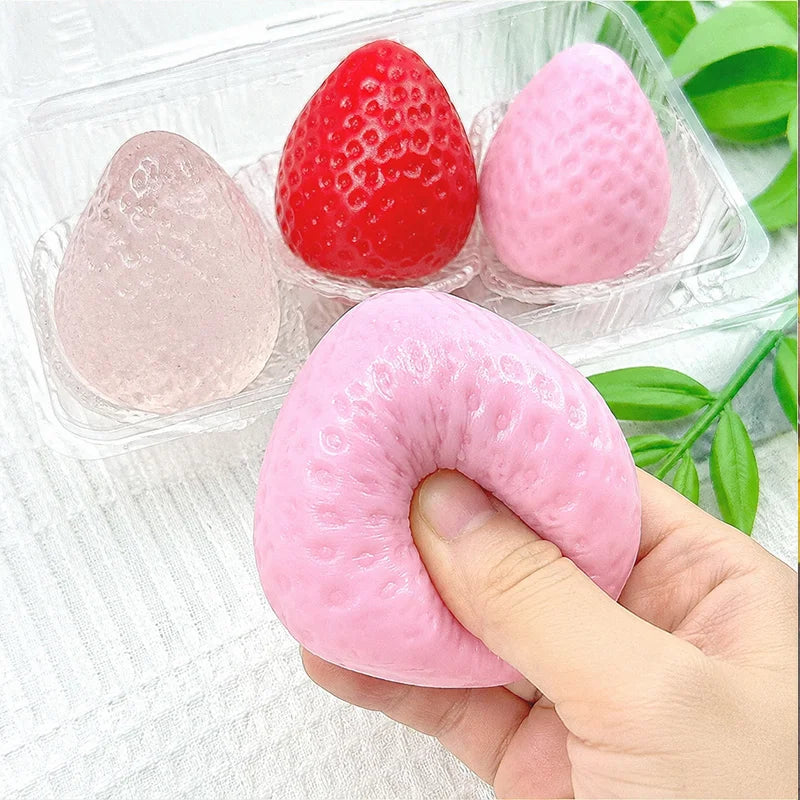 A hand holds a pink strawberry squishy toy next to two plastic containers, one containing a red and a pink strawberry squishy