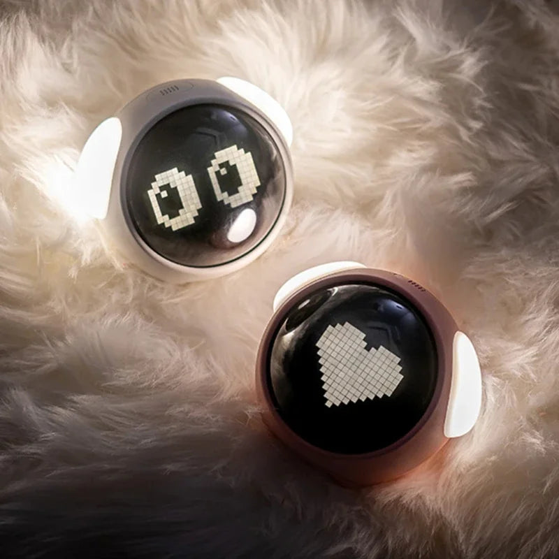 A white emoji alarm clock with a glowing display rests on a fluffy white surface.