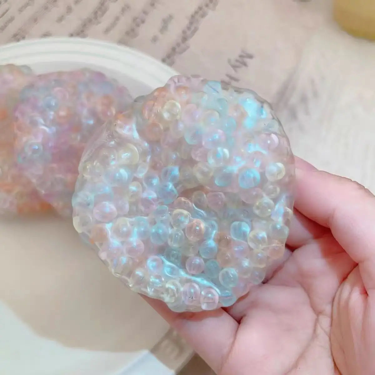 A hand holds a translucent, bubbly squishy stress relief ball with pink and blue iridescent colors.