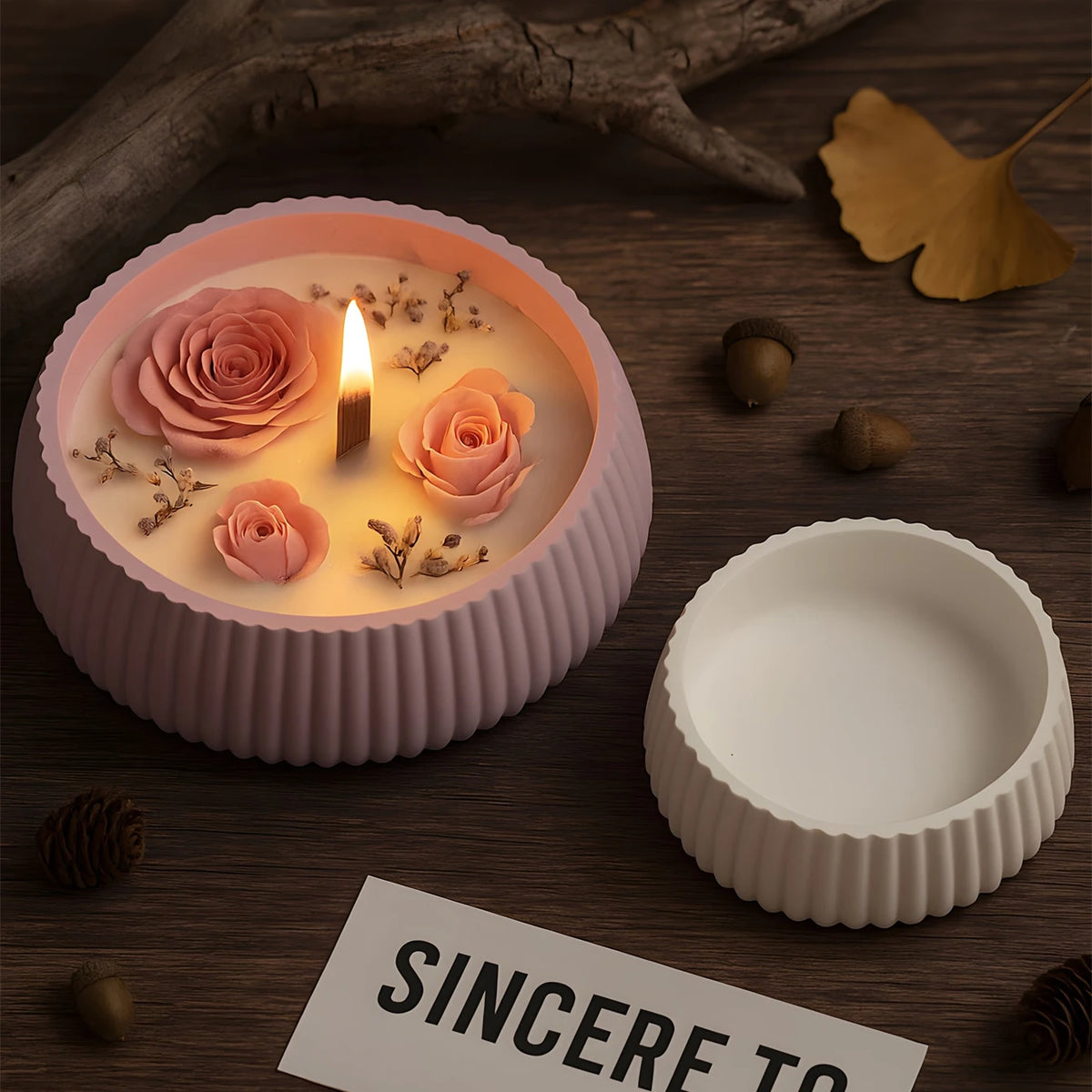 A pink candle in a round silicone mold sits next to a white circular mold and some dried floral elements on a wooden surface.