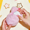 A hand holds a pink fluffy toy with a white earbud case cover and a gold hoop.