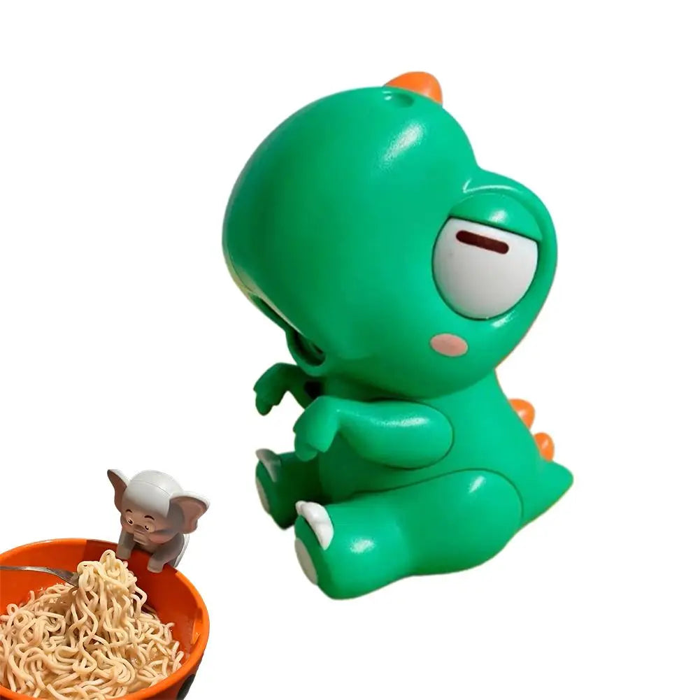 A small, green, plush toy resembling an elephant is shown next to a bowl of noodles.