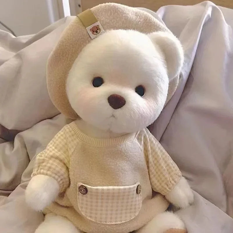 A white teddy bear plush wearing a cream-colored dress and a light beige hat, sitting on white bedding.