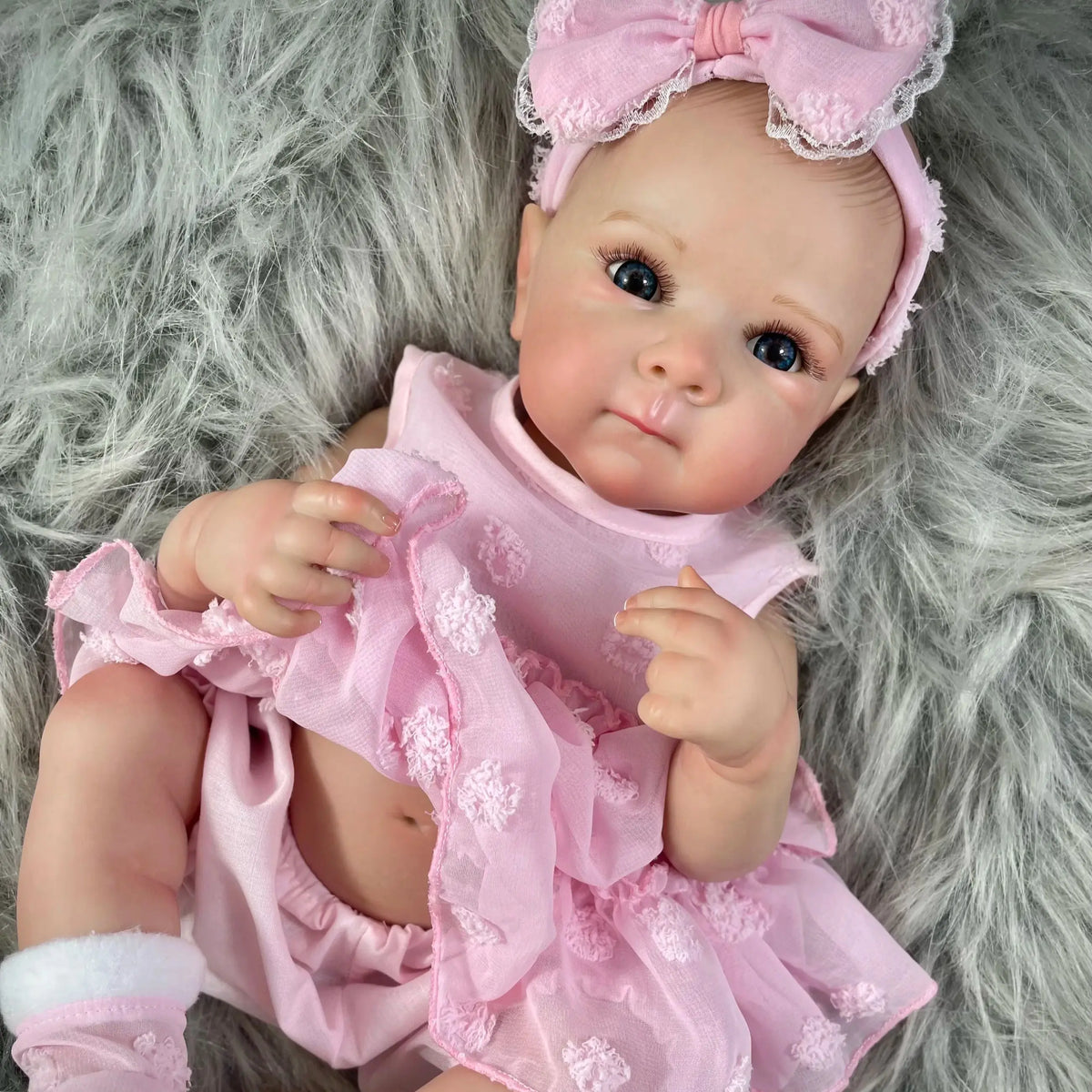 A pink reborn baby doll with painted hair wearing a ruffled dress is lying on a gray furry surface.