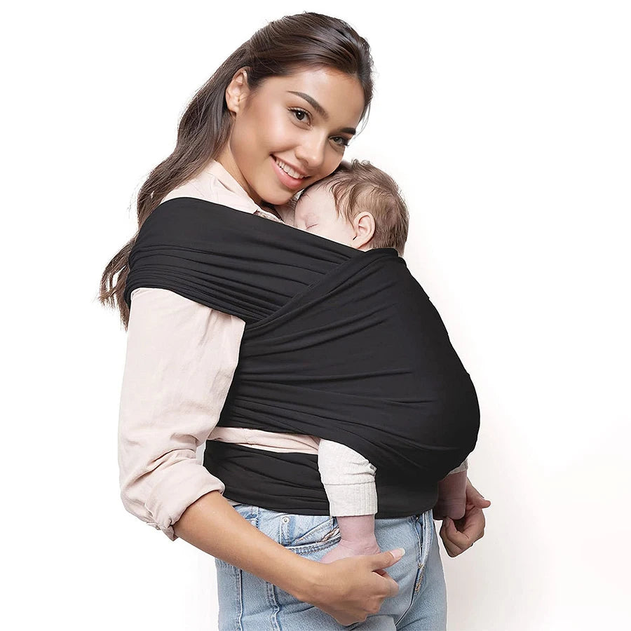 A woman wearing a black knitted fabric multi-functional baby carrier while smiling and holding a baby.