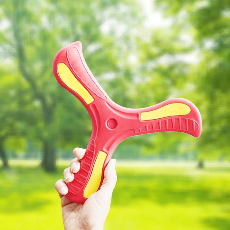 A hand holds a bright red soft foam boomerang flying disc with yellow accents in a green outdoor setting.