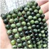 A hand holds a string of small, round natural stone beads in various shades of green and black.