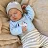 A newborn baby doll wearing a light blue long-sleeved shirt and striped pants is lying down.