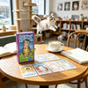 A box of Cat Tarot cards resting on a wooden table with a cup of coffee and a notebook.