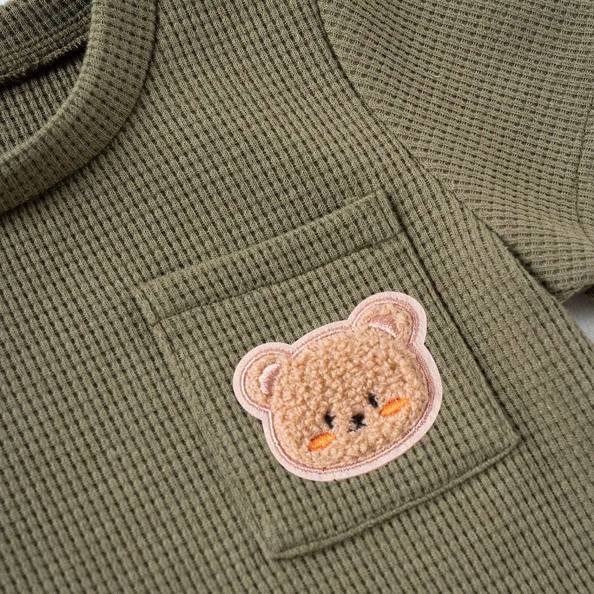 A close-up of a olive green knitted outfit featuring a cute, light brown bear print on the chest pocket.
