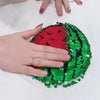 A hand holds a green and red sequin watermelon-shaped fabric against a white shirt.