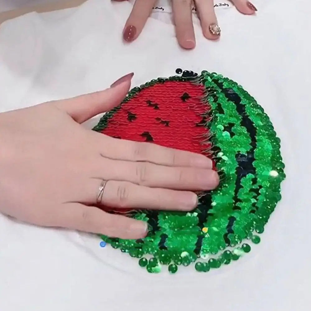 A hand holds a green and red sequin watermelon-shaped fabric against a white shirt.