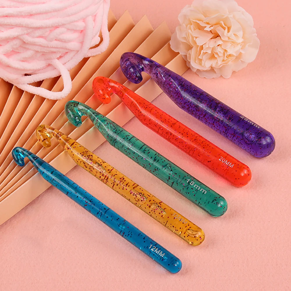 A set of three transparent crystal crochet hooks in purple, orange, and teal are displayed on a pink background.