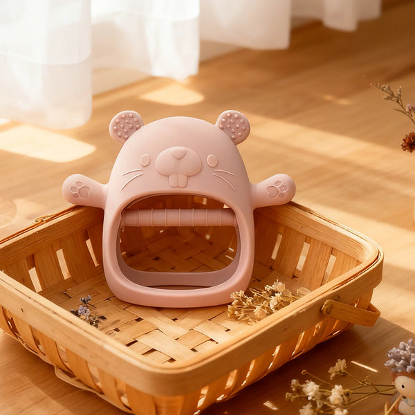 A pink silicone teething mitten shaped like a bear sits in a wooden basket with dried flowers.