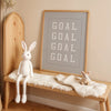 A framed canvas wall art featuring the word GOAL repeated in white text on a gray background, displayed on a wooden shelf wit