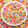 A white bowl filled with colorful polymer clay slice sprinkles for slime and DIY crafts scattered on a surface.