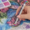 A hand holds a diamond painting pen with an interchangeable resin tip, resting on a sheet of diamond painting paper.