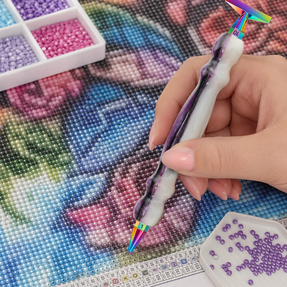 A hand holds a diamond painting pen with an interchangeable resin tip, resting on a sheet of diamond painting paper.