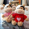 A plush cow stuffed animal with white and brown markings wearing a red sweater with a heart design.