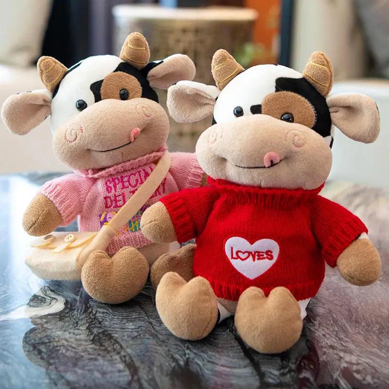 A plush cow stuffed animal with white and brown markings wearing a red sweater with a heart design.