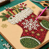 A hand is painting a Christmas tree design on a canvas using red, green, and white pearls and gems.