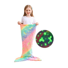 A young girl is holding a bright, glow-in-the-dark mermaid tail blanket with colorful scales and a dark background pattern.