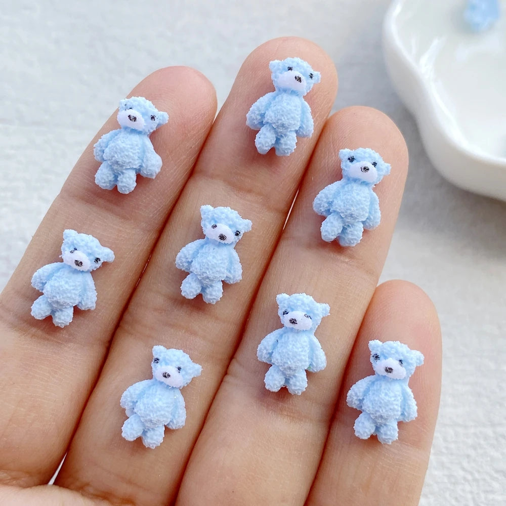 A hand displays several small, light blue resin teddy bear charms for DIY crafts.