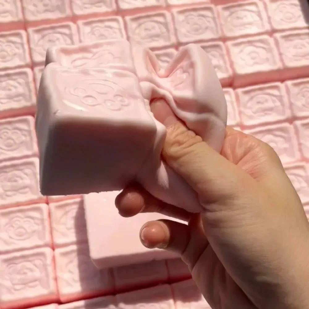 A hand holds a pink strawberry milk squishy stress relief toy against a pink grid background.