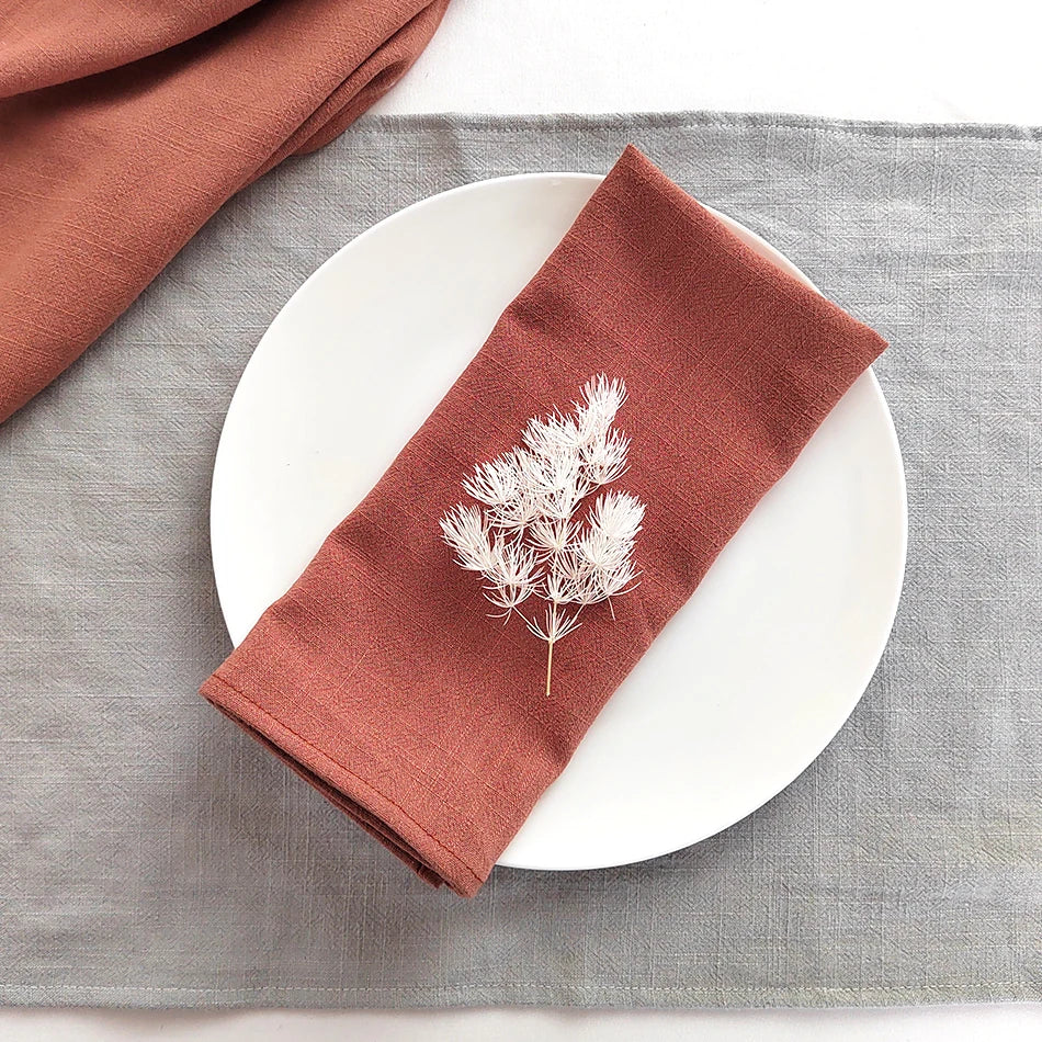 A folded terracotta colored linen napkin with a sprig of dried foliage placed on a white plate and gray linen cloth.