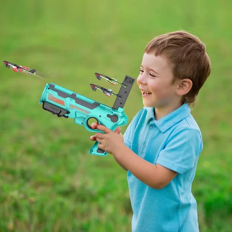 A young boy holds a teal airplane launcher toy with three foam planes in a grassy outdoor setting.