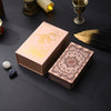 A box of gold foil tarot cards with a rose pink oracle finish resting on a dark surface.