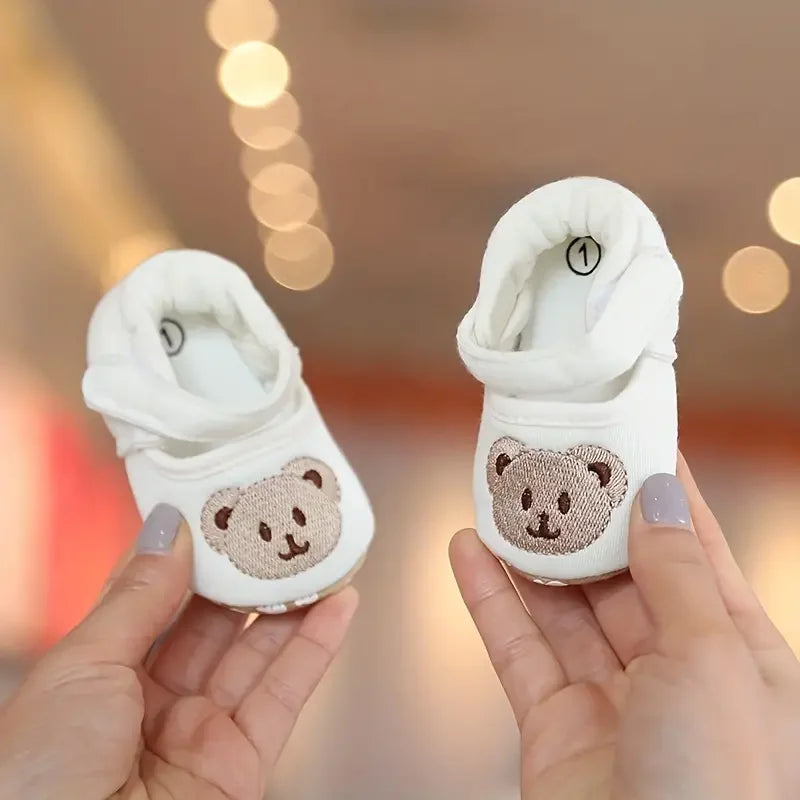 A pair of white soft sole first walker shoes featuring a cute bear design, held in a person's hands.