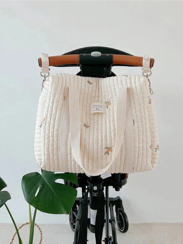 A cream quilted diaper bag with embroidery hangs on a black stroller frame.
