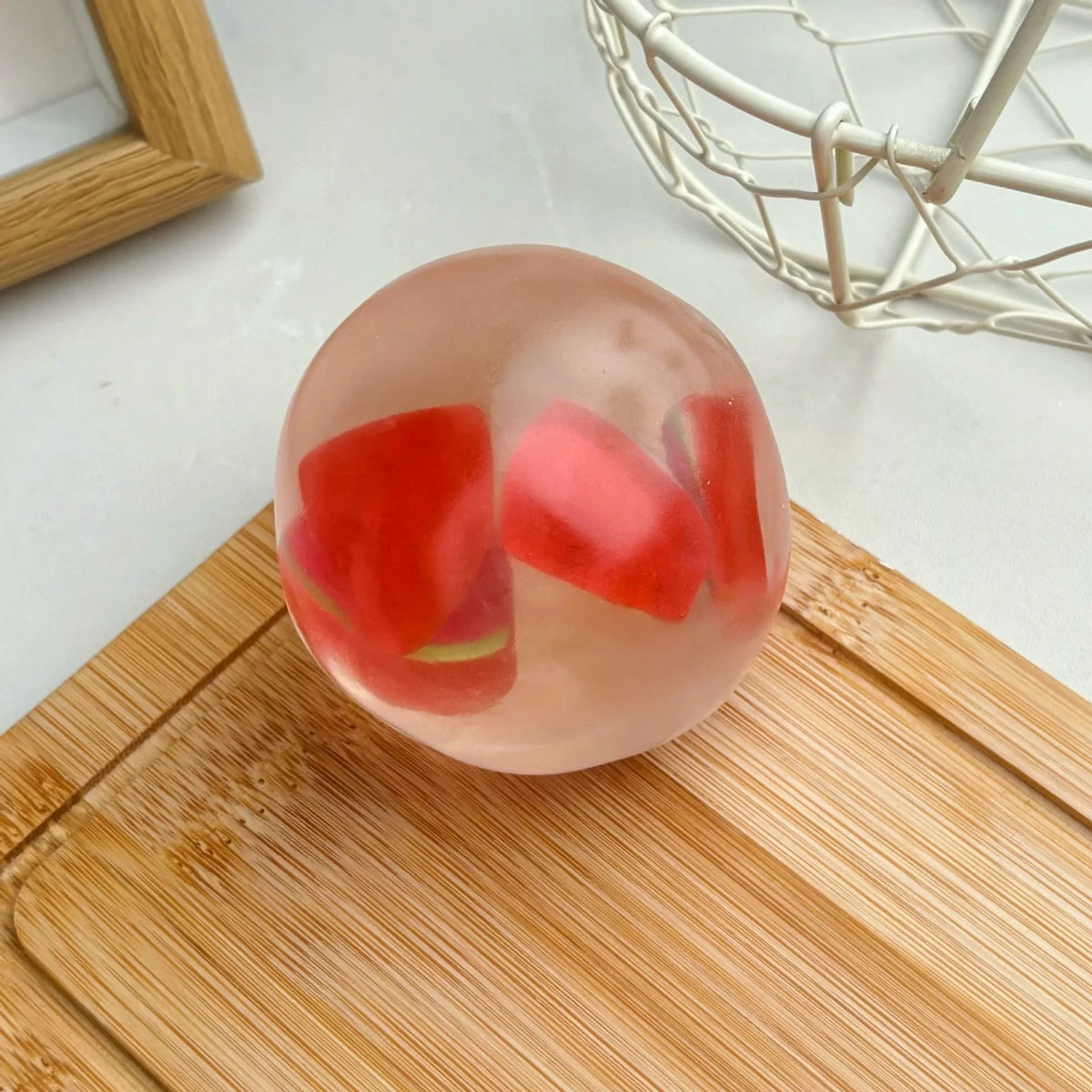 A clear pink squishy stress ball filled with red fruit shapes resting on a wooden cutting board.