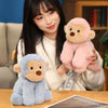 A woman holds a pink plush monkey soft toy and a blue plush monkey soft toy on a wooden table.