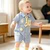 A baby boy wearing a blue short-sleeve romper with bear and mushroom appliqués standing on a rug.