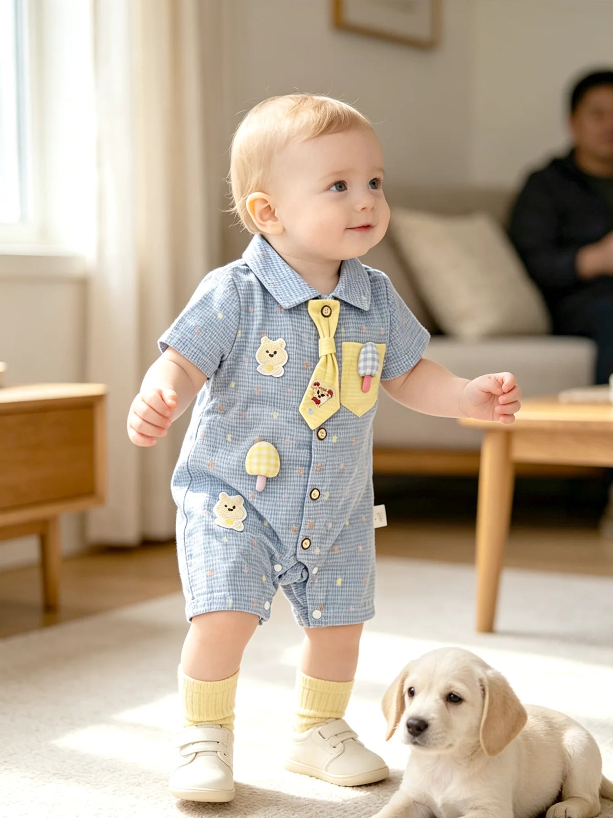 A baby boy wearing a blue short-sleeve romper with bear and mushroom appliqués standing on a rug.