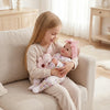A young girl is holding a realistic reborn baby doll with pink details on a light-colored sofa.