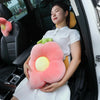 A woman is sitting in a car, holding a large pink plush pillow shaped like a flower with yellow accents.