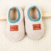 Pair of white plush baby slippers with light blue trim and orange WLS tags resting on a cream towel.