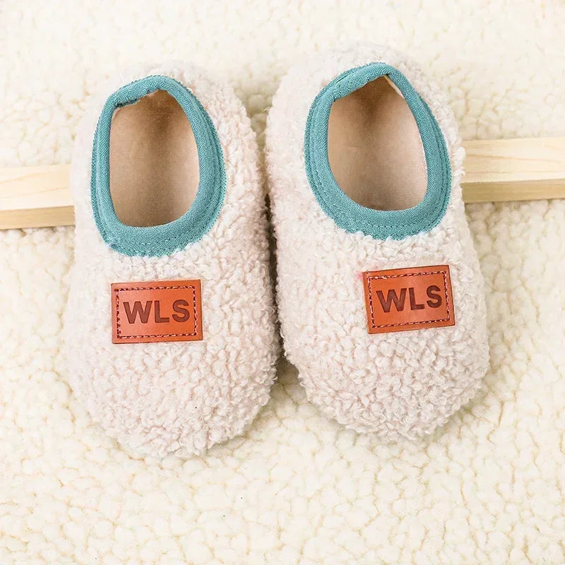 Pair of white plush baby slippers with light blue trim and orange WLS tags resting on a cream towel.