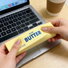 A hand holds a yellow salted butter squishy toy next to a laptop.