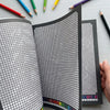 A close-up view of a book with black and white pixel art mosaics for adults, surrounded by colored pencils.