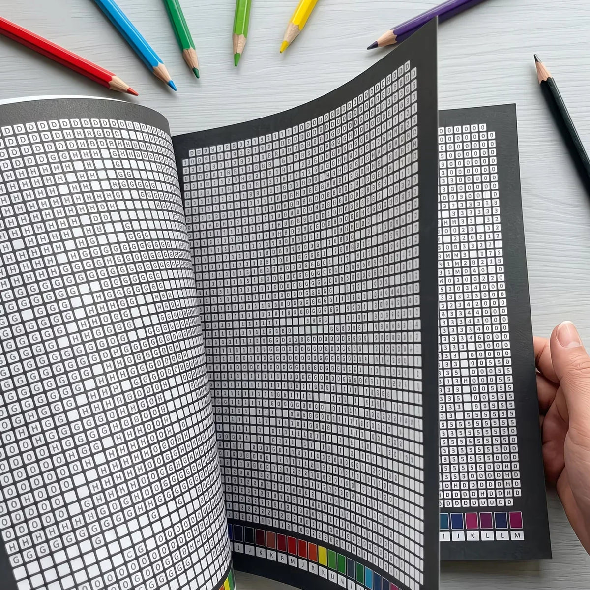 A close-up view of a book with black and white pixel art mosaics for adults, surrounded by colored pencils.