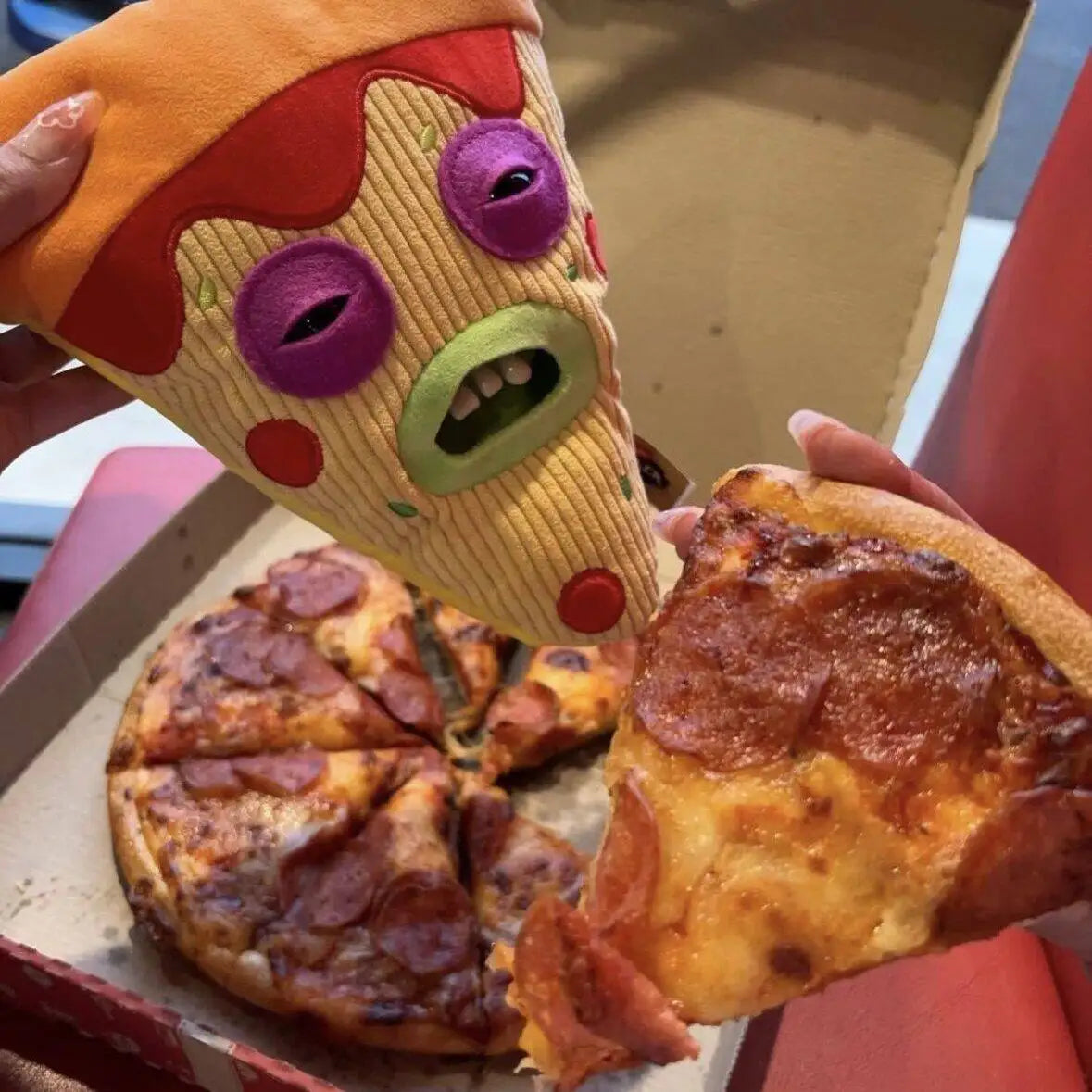 A hand holds a slice of pizza and a plush toy shaped like an ugly cute monster.