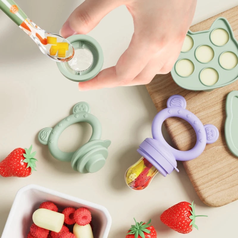 A hand is using a silicone baby fruit feeder with a yellow-green fruit to feed a baby, surrounded by various silicone fruit m